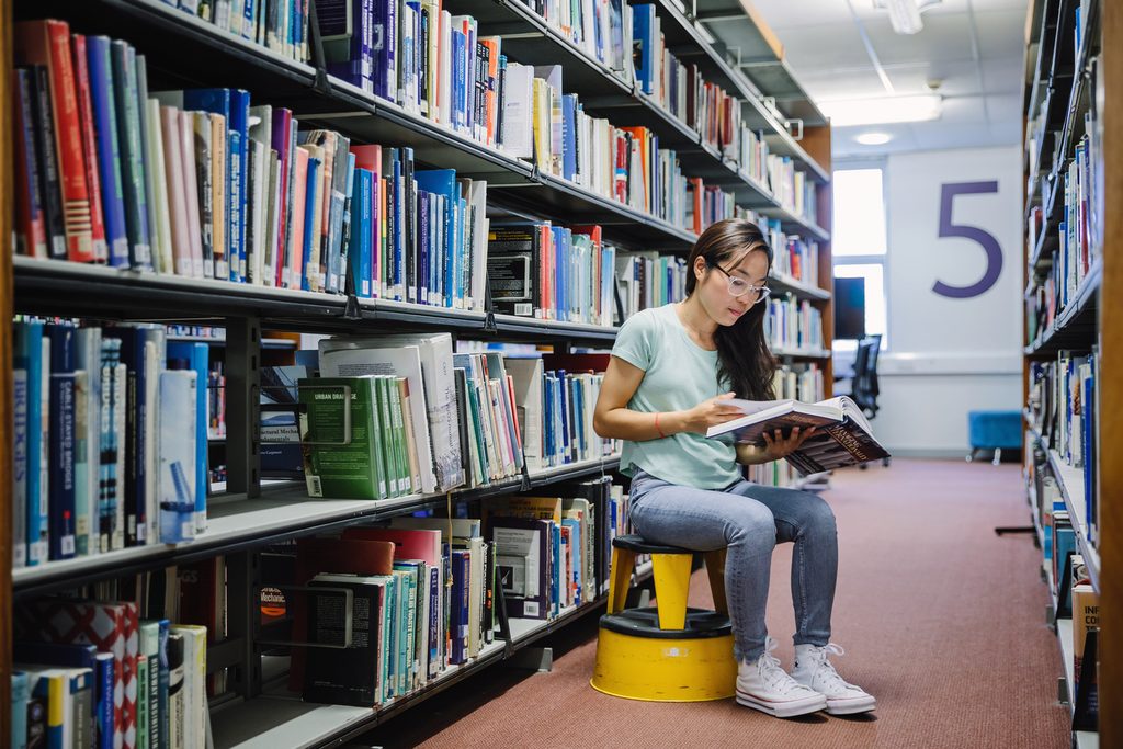 Student sitting in front of the book shelves and reading a book in the library